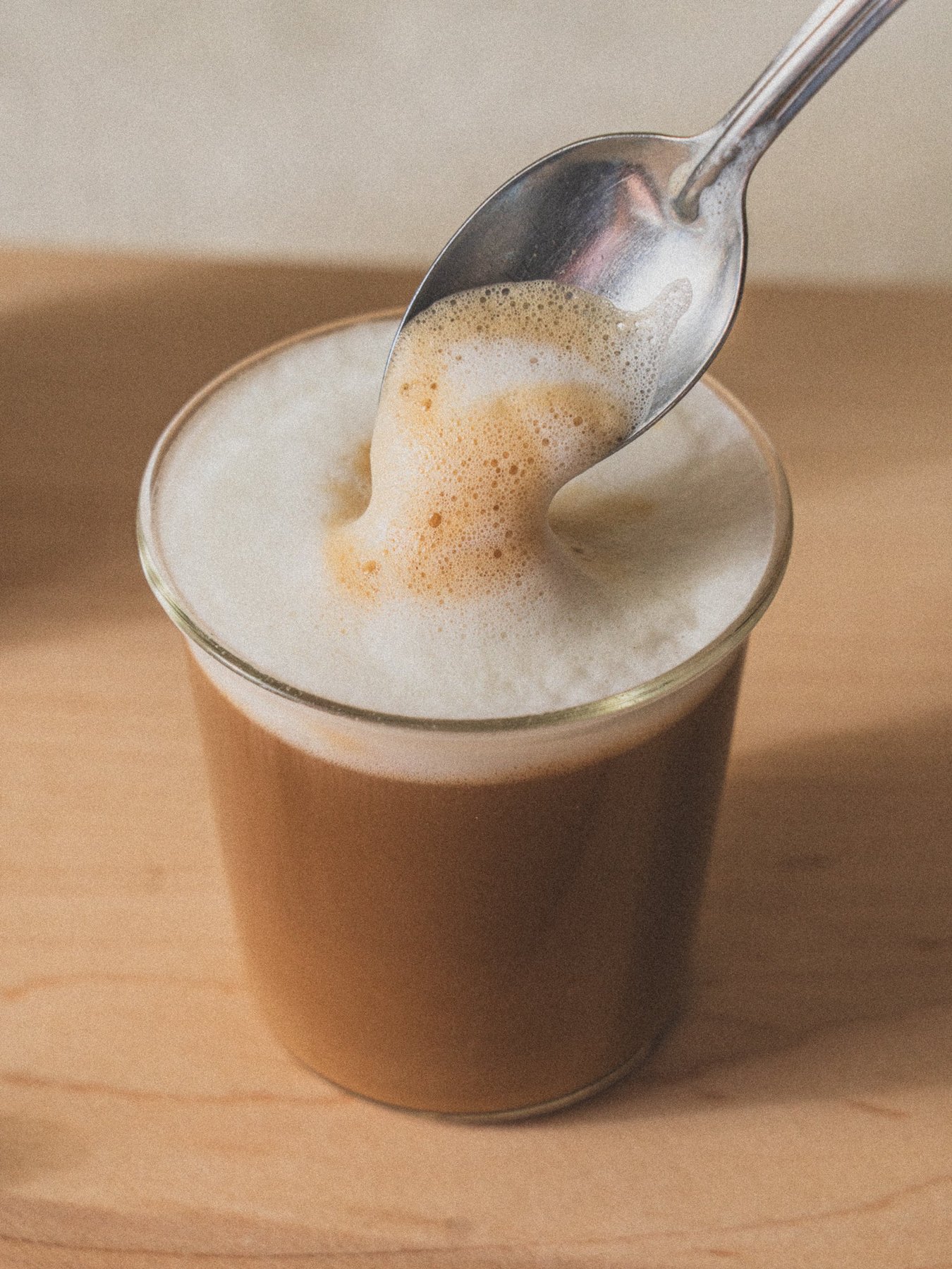 Foamy latte in a clear glass with a spoon dripping foam above.