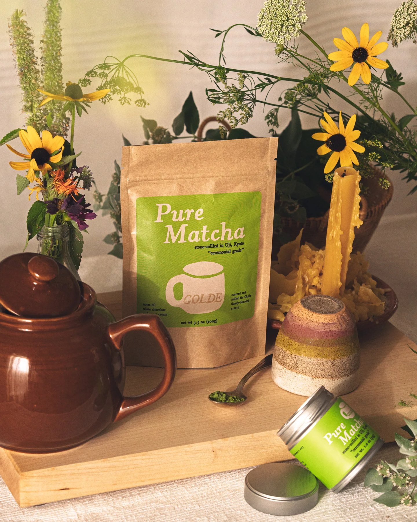 Matcha package displayed beside brown teapot candles blooms and spice tin.