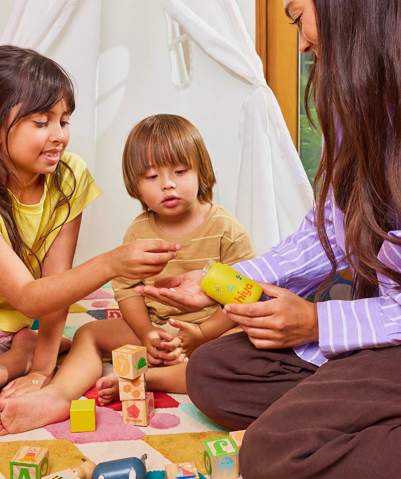 A woman offers a yellow Hiya bottle to two small children playing.