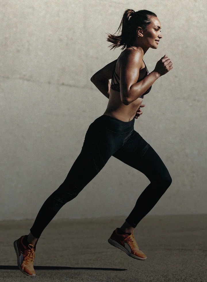 Athletic woman sprinting outdoors in black leggings and orange shoes.