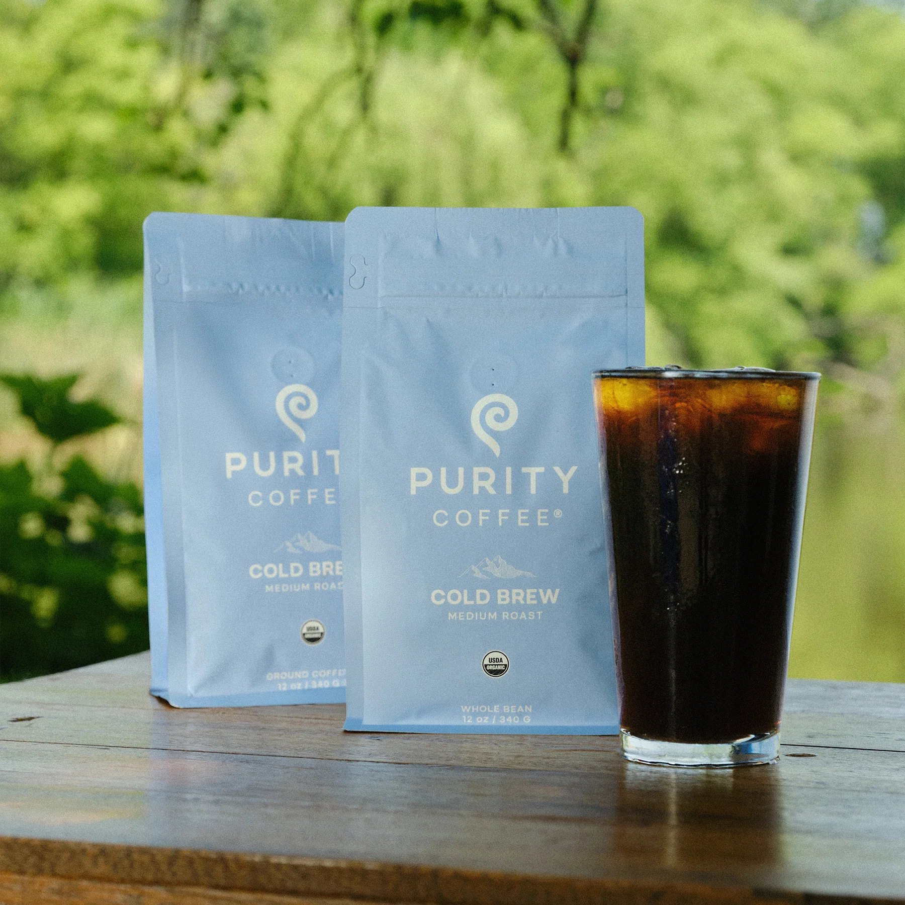 Two blue Cold Brew bags stand beside iced coffee outdoors.